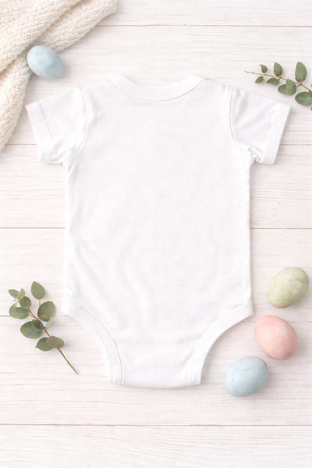 White baby onesie on a light wooden surface with pastel-colored eggs and greenery showing back of bodysuit.